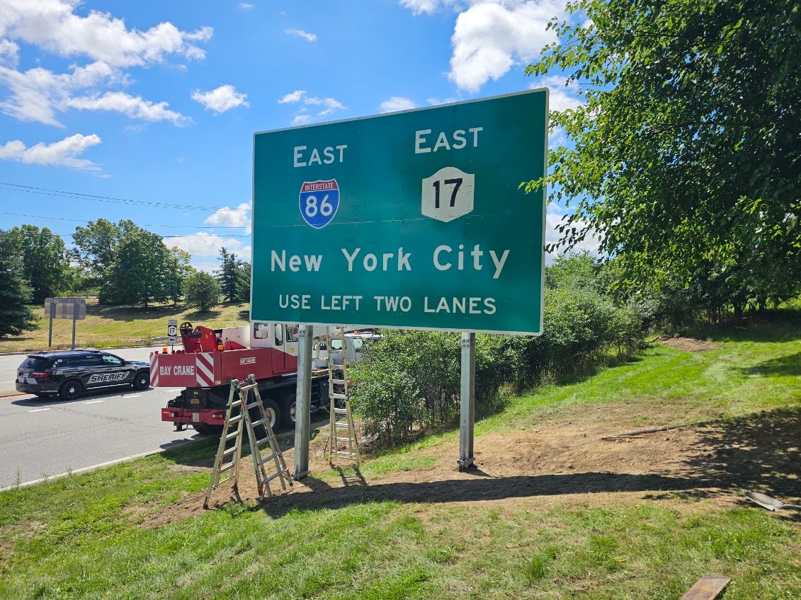 Crew installing a large I-86 and Route 17 East guide sign to New York City on the highway shoulder with a boom truck and police escort